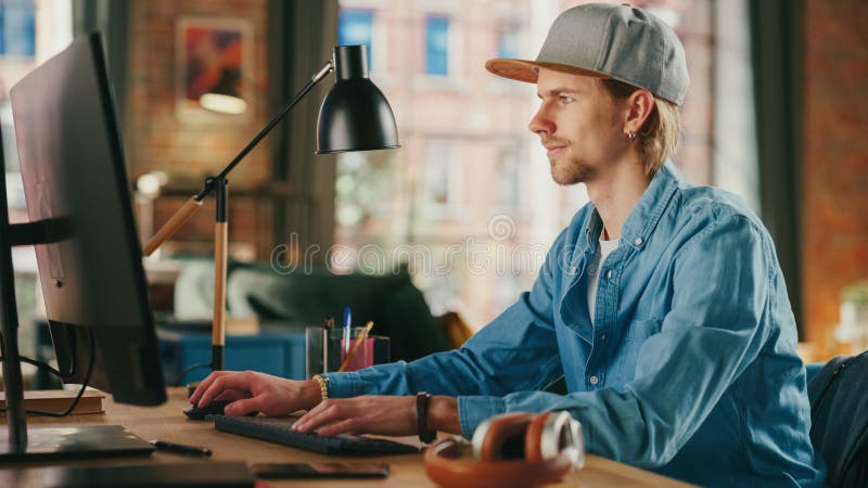 Young Handsome Man Working from Loft Apartment on Desktop Computer. Creative Male Checking Social Media, Browsing Internet from Home. Urban City View from Big Window. Young Handsome Man Working from Loft Apartment on Desktop Computer. Creative Male Checking Social Media, Browsing Internet from Home. Urban City View from Big Window.