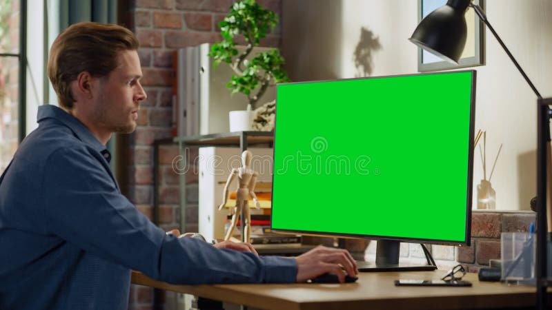 Young Handsome Man Working from Home on Desktop Computer with Green Screen Mock Up Display. Male Checking Corporate Accounts, Messaging Colleagues. Loft Living Room with Big Window. Young Handsome Man Working from Home on Desktop Computer with Green Screen Mock Up Display. Male Checking Corporate Accounts, Messaging Colleagues. Loft Living Room with Big Window.