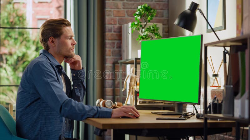 Young Handsome Man Working from Home on Desktop Computer with Green Screen Mock Up Display. Male Checking Corporate Accounts, Messaging Colleagues. Loft Living Room with Big Window. Young Handsome Man Working from Home on Desktop Computer with Green Screen Mock Up Display. Male Checking Corporate Accounts, Messaging Colleagues. Loft Living Room with Big Window.