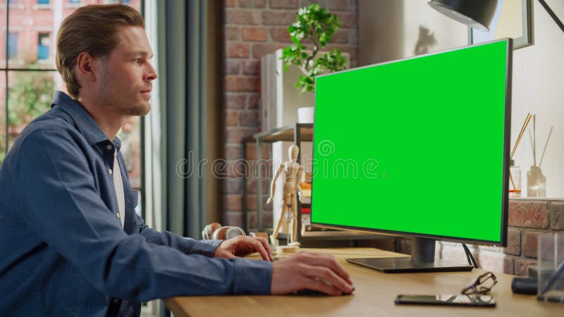 Young Handsome Man Working from Home on Desktop Computer with Green Screen Mock Up Display. Male Checking Corporate Accounts, Messaging Colleagues. Loft Living Room with Big Window. Young Handsome Man Working from Home on Desktop Computer with Green Screen Mock Up Display. Male Checking Corporate Accounts, Messaging Colleagues. Loft Living Room with Big Window.