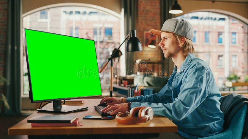 Young Handsome Man Working from Home on Desktop Computer with Green Screen Mock Up Display. Student Making Homework Assignment, Studying for Exams. Urban City View from Big Window. Young Handsome Man Working from Home on Desktop Computer with Green Screen Mock Up Display. Student Making Homework Assignment, Studying for Exams. Urban City View from Big Window.