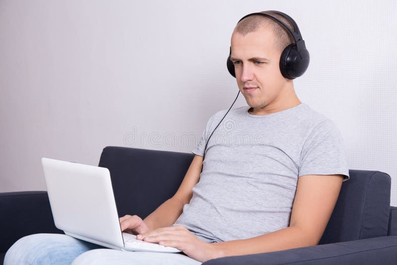 Jeune homme bel s'asseyant sur le sofa avec l'ordinateur et le m de écoute images libres de droits Jeune homme bel s'asseyant sur le sofa avec l'ordinateur et le m de écoute images libres de droits