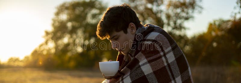 Jeune homme congelé dans le thé potable couvrant chaud se reposant dans le domaine pendant le sunet f photos stock Jeune homme congelé dans le thé potable couvrant chaud se reposant dans le domaine pendant le sunet f photos stock