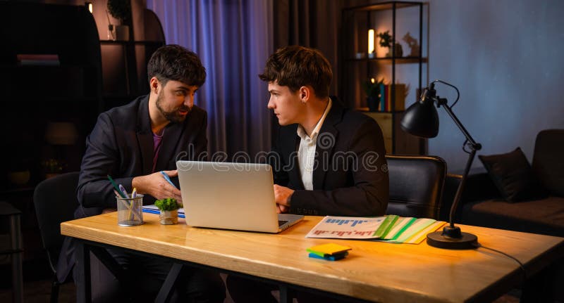 Jeune homme d'affaires au bureau qui travaille tard le soir assis au bureau avec un ordinateur portable parlant avec un assistant photos libres de droits