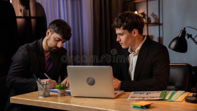 Jeune homme d'affaires au bureau qui travaille tard le soir assis au bureau avec un ordinateur portable parlant avec un assistant image libre de droits