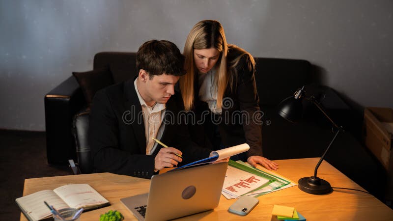 Jeune homme d'affaires au bureau qui travaille tard le soir assis au bureau avec un ordinateur portable parlant avec une assistant images libres de droits