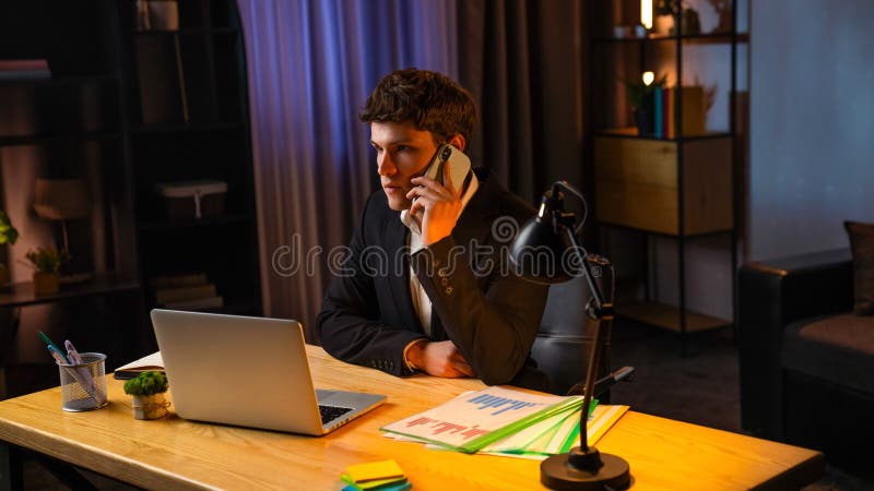 Jeune homme d'affaires dans le bureau travaillant tard le soir homme assis au bureau utilisant un ordinateur portable parlant sur photos stock