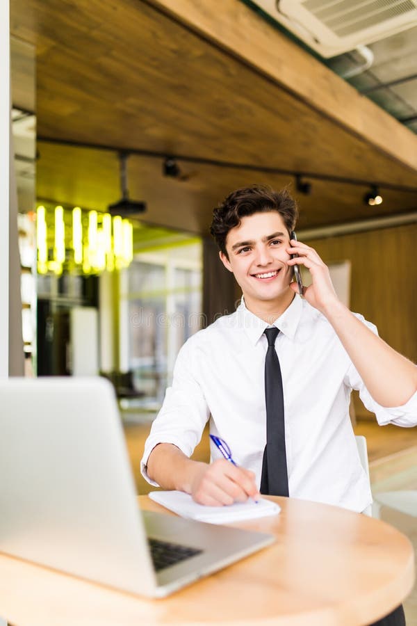 Jeune homme d'affaires de sourire s'asseyant derrière son bureau avec l'ordinateur portable et parlant au téléphone portable dans photo stock Jeune homme d'affaires de sourire s'asseyant derrière son bureau avec l'ordinateur portable et parlant au téléphone portable dans photo stock