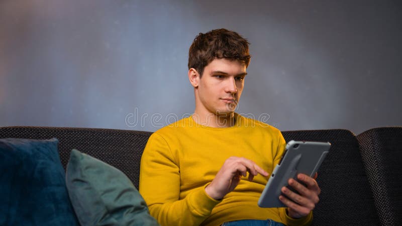 Jeune homme dans la soirée après le travail se détendre à la maison assis sur le canapé dans le salon regarder la vidéo sur la tab photo stock
