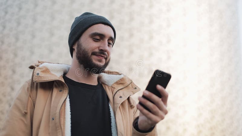 Jeune homme de barbe envoyant le message audio sur le téléphone portable à extérieur Il utilise une veste d'automne et un chapeau banque de vidéos