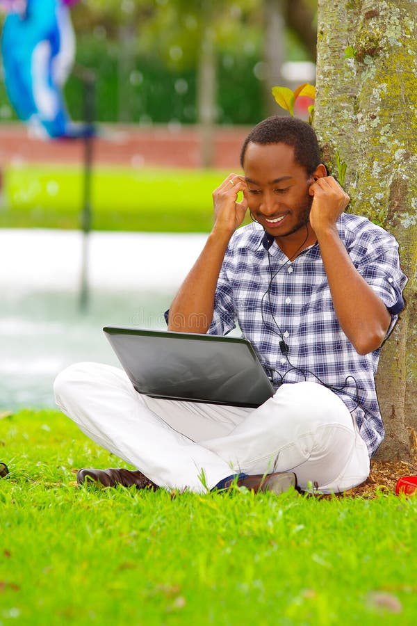 Jeune homme de couleur s'asseyant sur l'herbe verte et travaillant dans son ordinateur et musique de écoute avec sa pose d'écoute image libre de droits Jeune homme de couleur s'asseyant sur l'herbe verte et travaillant dans son ordinateur et musique de écoute avec sa pose d'écoute image libre de droits