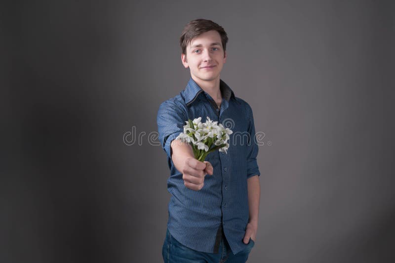 Jeune homme de sourire beau dans la chemise bleue se tenant dans le bouquet tendu de main avec les perce-neige blancs et regardan photographie stock libre de droits Jeune homme de sourire beau dans la chemise bleue se tenant dans le bouquet tendu de main avec les perce-neige blancs et regardan photographie stock libre de droits