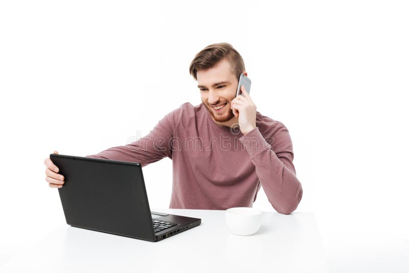 Jeune homme de sourire regardant l'affichage de l'ordinateur parlant au téléphone et jugeant l'affichage d'ordinateur portable d' image stock Jeune homme de sourire regardant l'affichage de l'ordinateur parlant au téléphone et jugeant l'affichage d'ordinateur portable d' image stock