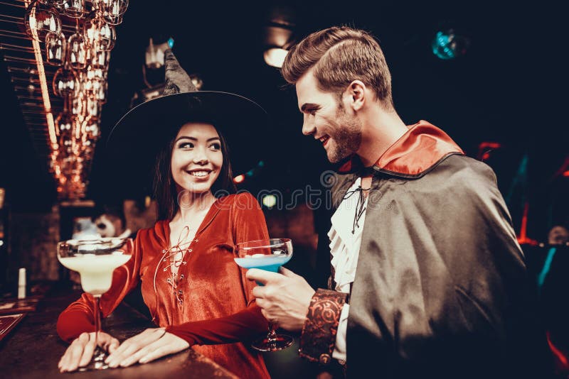 Young Man and Woman in Costumes at Halloween Party. Handsome Man and Beautiful Woman Wearing Halloween Costumes Standing next to Bar counter in Nightclub. Celebration of Halloween. Young Man and Woman in Costumes at Halloween Party. Handsome Man and Beautiful Woman Wearing Halloween Costumes Standing next to Bar counter in Nightclub. Celebration of Halloween