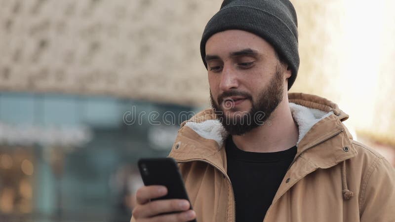 Jeune homme heureux de barbe utilisant le smartphone dans la rue près du centre commercial Il utilise une veste d'automne et un c banque de vidéos