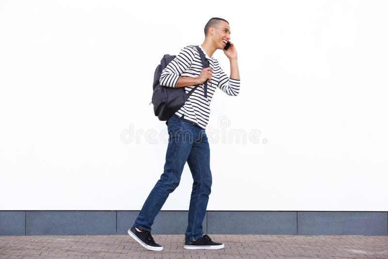 Jeune homme heureux marchant avec le sac et parlant au téléphone portable contre le mur blanc image stock Jeune homme heureux marchant avec le sac et parlant au téléphone portable contre le mur blanc image stock