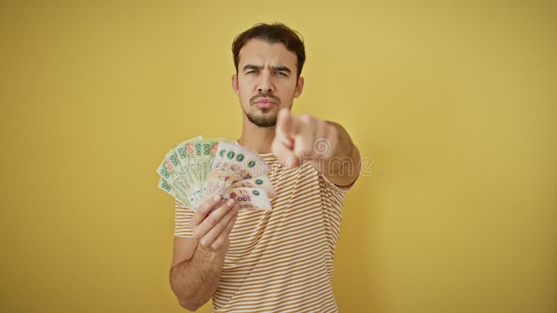 Jeune homme hispanique confiant vous pointe en tenant de l'argent en faisant un geste sérieux et direct sur un jaune isolé banque de vidéos