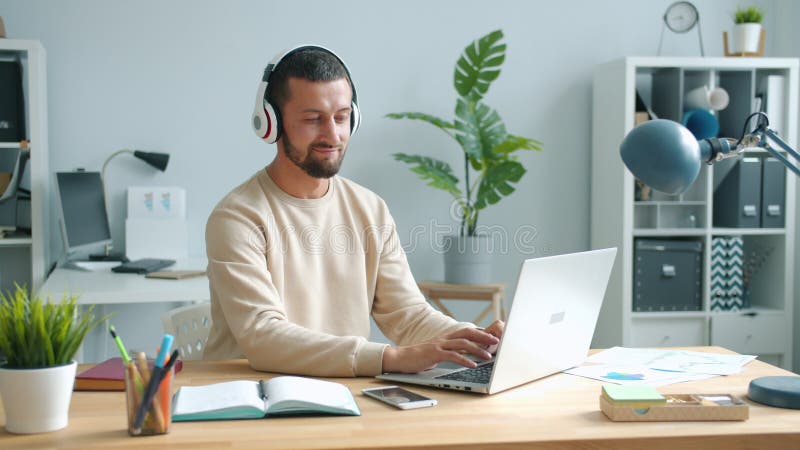 Jeune homme joyeux aux casques utilisant un ordinateur au bureau travaillant avec un ordinateur portable banque de vidéos