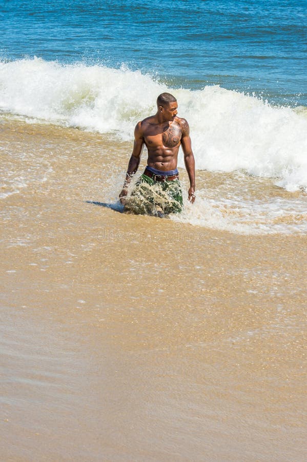 Jeune homme noir torse nu s'agenouillant sur la plage jouant sur des vagues d'eau photos stock