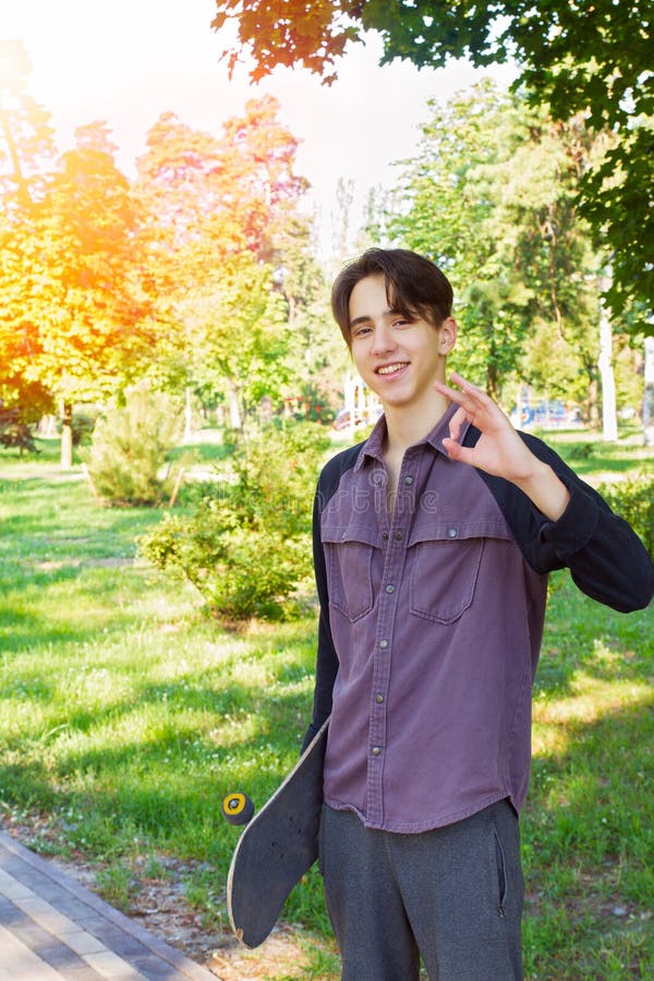 Jeune homme se tenant avec la planche à roulettes dans des mains en parc de ville Patineur de l'adolescence de garçon image stock Jeune homme se tenant avec la planche à roulettes dans des mains en parc de ville Patineur de l'adolescence de garçon image stock