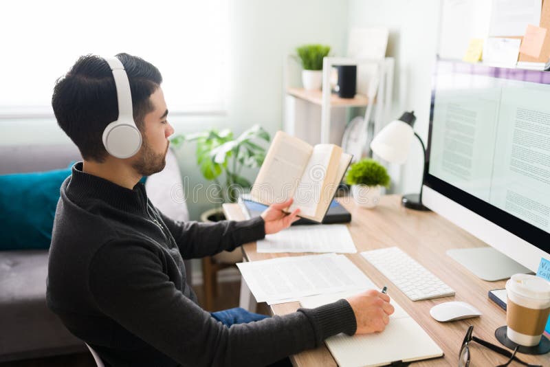 Man in his 20s sitting at his desk at the office and listening to music with headphones. Translator writing and working with a notebook a. Man in his 20s sitting at his desk at the office and listening to music with headphones. Translator writing and working with a notebook a