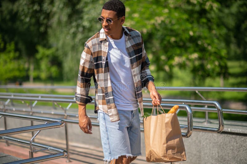 Jeune homme à la peau foncée en chemise plaid marchant avec un sac en papier dans les mains images libres de droits