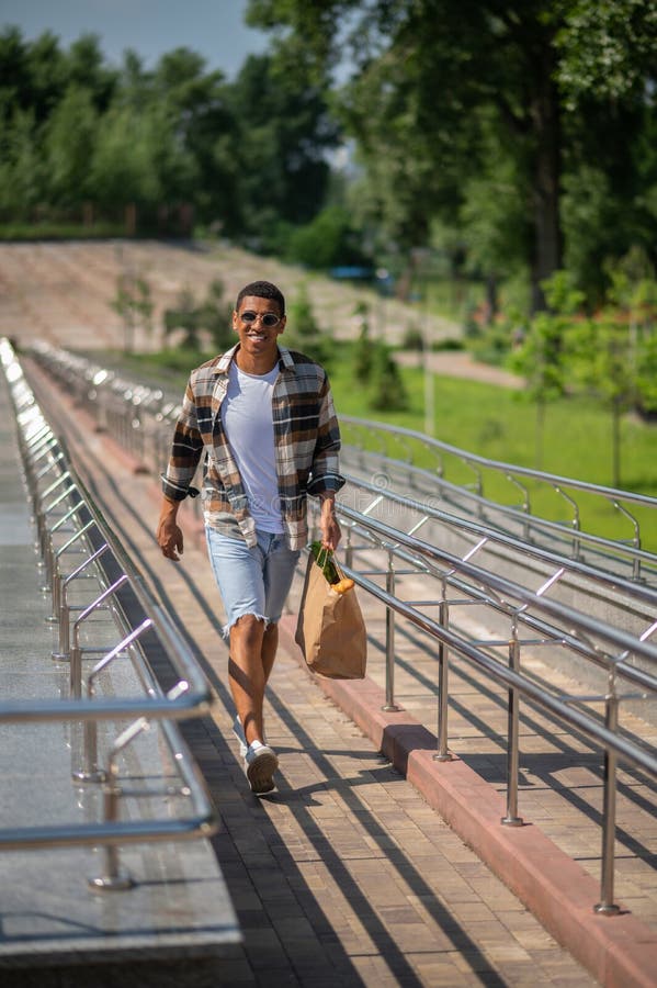 Jeune homme à la peau foncée en chemise plaid marchant avec un sac en papier dans les mains images libres de droits