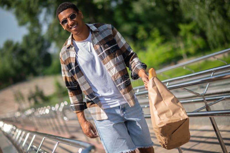 Jeune homme à la peau foncée en chemise plaid marchant avec un sac en papier dans les mains images libres de droits