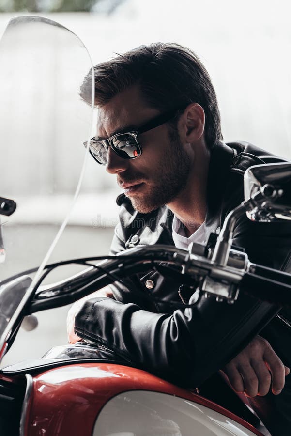 Jeune homme élégant beau dans les lunettes de soleil et la veste en cuir se reposant sur la moto photo stock Jeune homme élégant beau dans les lunettes de soleil et la veste en cuir se reposant sur la moto photo stock