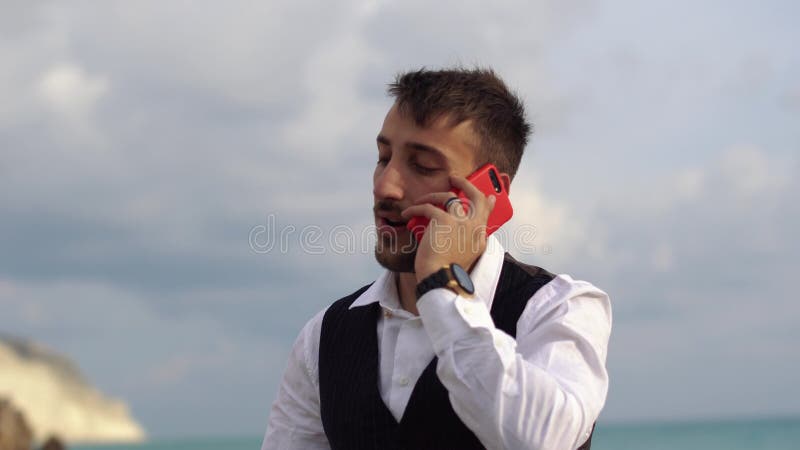 Jeune homme élégant mignon parlant par la cellule près de la vue de côté stupéfiante de mer Homme d'affaires parlant par le télép banque de vidéos