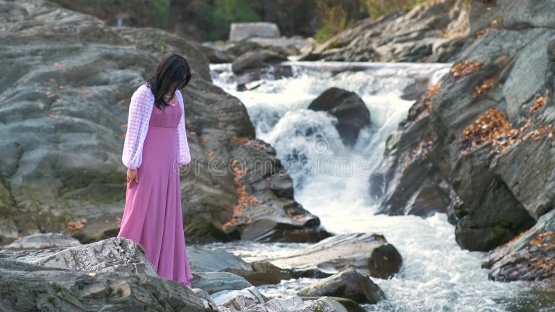 Jeune jolie femme en longue robe de soirée à la mode debout près de la petite rivière de montagne avec l'eau en mouvement rapide. banque de vidéos