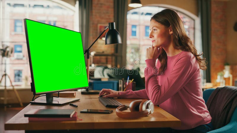 Young Pretty Female Working from Home on Desktop Computer with Green Screen Mock Up Display. Creative Woman Checking Social Media, Browsing Internet. Loft Apartment with City View from Big Window. Young Pretty Female Working from Home on Desktop Computer with Green Screen Mock Up Display. Creative Woman Checking Social Media, Browsing Internet. Loft Apartment with City View from Big Window.