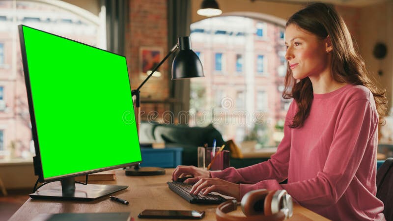 Young Pretty Female Working from Home on Desktop Computer with Green Screen Mock Up Display. Creative Woman Checking Social Media, Browsing Internet. Loft Apartment with City View from Big Window. Young Pretty Female Working from Home on Desktop Computer with Green Screen Mock Up Display. Creative Woman Checking Social Media, Browsing Internet. Loft Apartment with City View from Big Window.