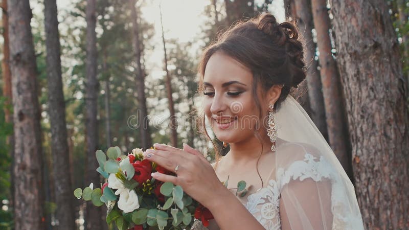 Jeune mariée avec du charme et heureuse dans une belle robe de mariage avec un bouquet des fleurs dans une forêt de pin au soleil