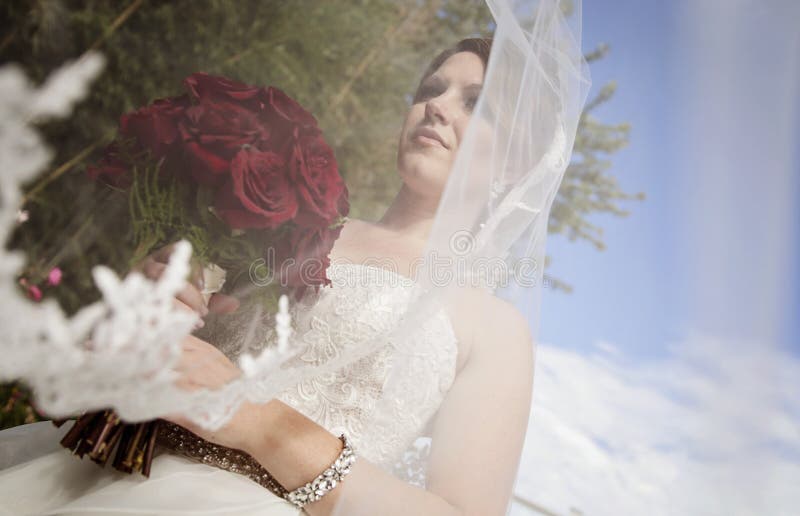 Jeune mariée avec le long voile soufflant en vent. Un profil bas d'une jeune mariée avec le long voile soufflant dans le vent tenant un bouquet des roses rouges
