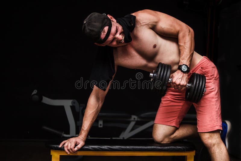 Jeune mâle adulte beau faisant des rangées d'haltère d'un-bras sur le banc dans le gymnase photo stock Jeune mâle adulte beau faisant des rangées d'haltère d'un-bras sur le banc dans le gymnase photo stock