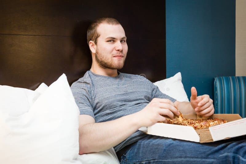 Jeune mâle caucasien s'asseyant sur le lit mangeant de la pizza photographie stock libre de droits Jeune mâle caucasien s'asseyant sur le lit mangeant de la pizza photographie stock libre de droits