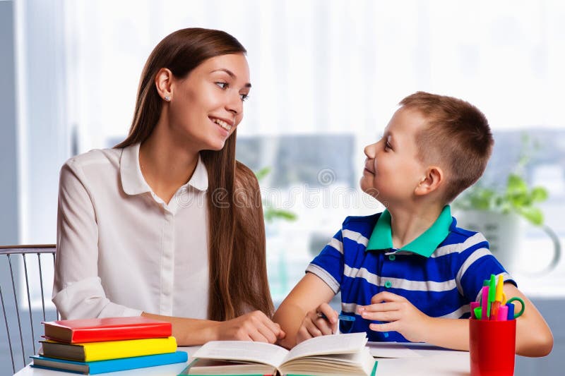 Jeune mère s'asseyant à une table à la maison aidant son petit fils avec ses devoirs d'école comme il écrit des notes dans un car images stock Jeune mère s'asseyant à une table à la maison aidant son petit fils avec ses devoirs d'école comme il écrit des notes dans un car images stock