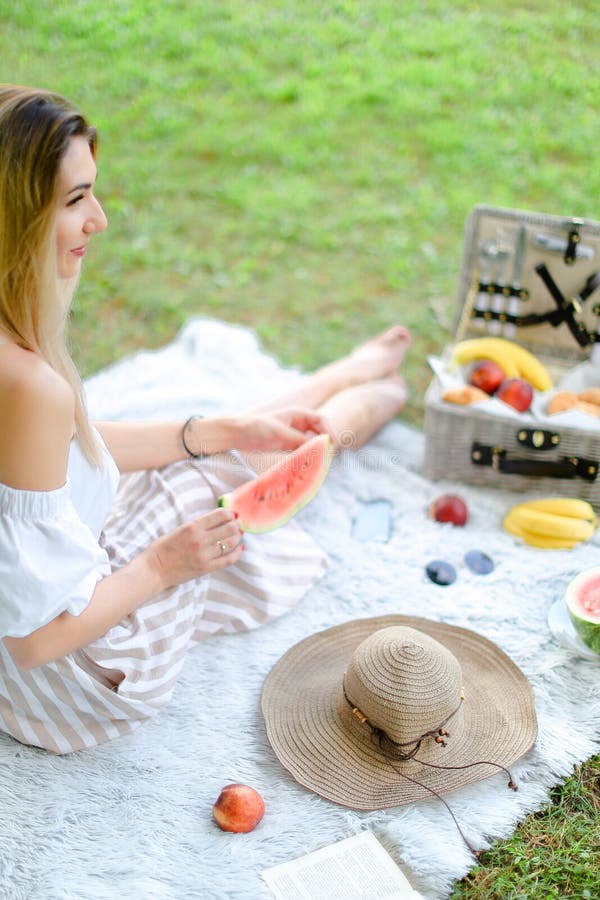Jeune personne féminine blonde s'asseyant sur le plaid près des fruits et du chapeau, mangeant la pastèque, herbe à l'arrière-pla photos libres de droits Jeune personne féminine blonde s'asseyant sur le plaid près des fruits et du chapeau, mangeant la pastèque, herbe à l'arrière-pla photos libres de droits