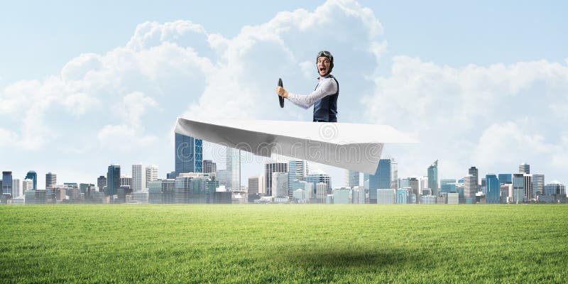 Young pilot sitting in big paper plane. Modern urban architecture with high skyscrapers on background. Man in paper airplane flying low above green field. Megalopolis panorama with green grass. Young pilot sitting in big paper plane. Modern urban architecture with high skyscrapers on background. Man in paper airplane flying low above green field. Megalopolis panorama with green grass