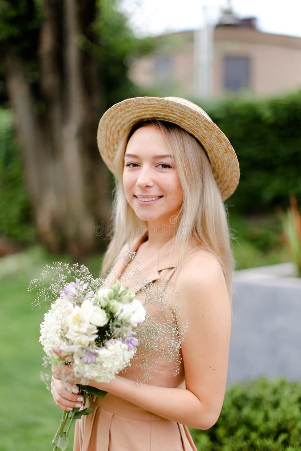 Jeune position caucasienne de fille dans le yeard avec le bouquet des fleurs et du chapeau de port images stock Jeune position caucasienne de fille dans le yeard avec le bouquet des fleurs et du chapeau de port images stock