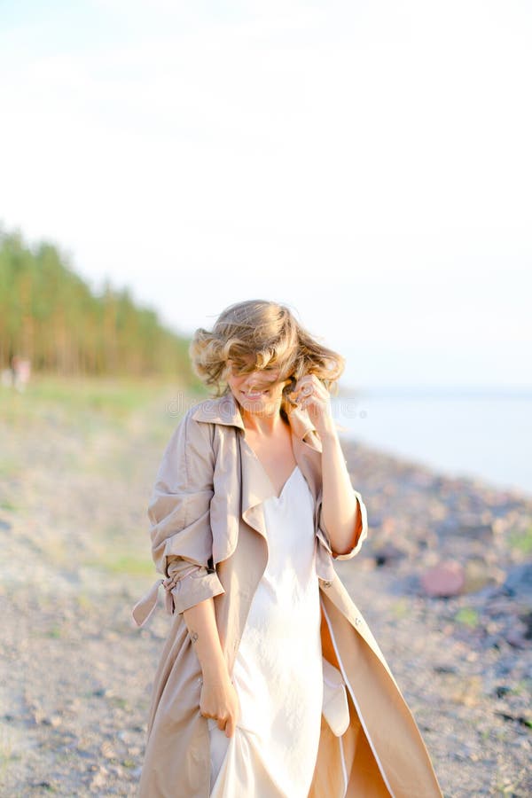 Jeune position de sourire de femme sur la plage de mer et le manteau de port avec la robe blanche photographie stock libre de droits Jeune position de sourire de femme sur la plage de mer et le manteau de port avec la robe blanche photographie stock libre de droits