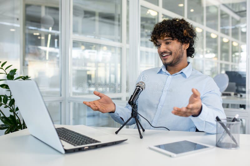 An enthusiastic young man in a modern office space is engaging with an online audience via microphone and laptop. He displays a lively expression and gestures. An enthusiastic young man in a modern office space is engaging with an online audience via microphone and laptop. He displays a lively expression and gestures.