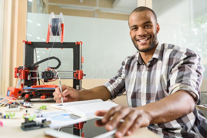 This 3d printer is wonderful. Joyful male engineer is doing research in technology lab. He is sitting and making notes. Man is looking at camera and smiling. This 3d printer is wonderful. Joyful male engineer is doing research in technology lab. He is sitting and making notes. Man is looking at camera and smiling