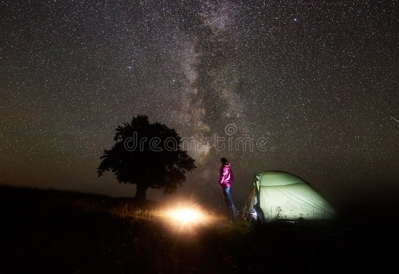 Night camping in mountains. Hiker woman standing near illuminated tent and silhouette of big tree, enjoying brightly burning campfire under beautiful dark starry sky and Milky way. Tourism concept. Night camping in mountains. Hiker woman standing near illuminated tent and silhouette of big tree, enjoying brightly burning campfire under beautiful dark starry sky and Milky way. Tourism concept