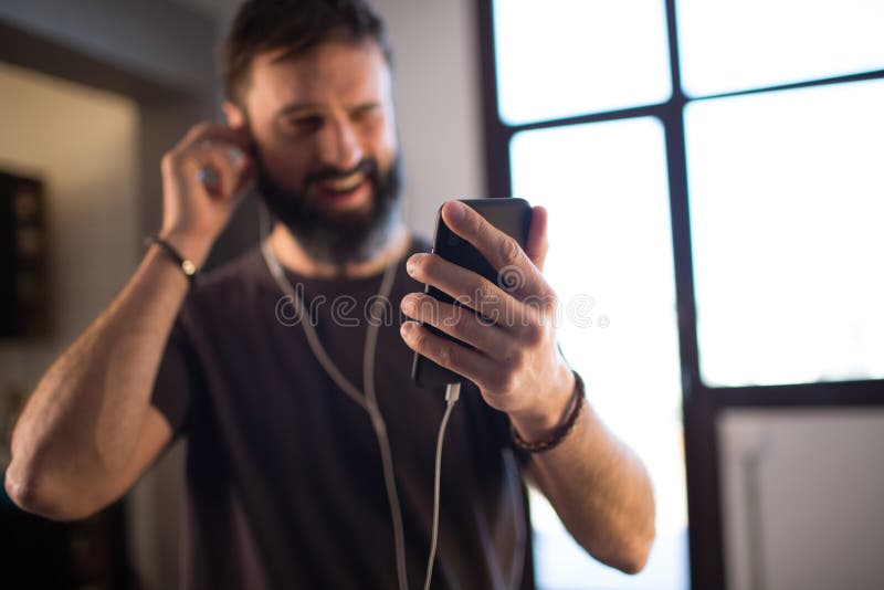 Jeune type barbu portant la musique de écoute de T-shirt gris occasionnel dans des écouteurs, vérifiant les réseaux sociaux s image libre de droits Jeune type barbu portant la musique de écoute de T-shirt gris occasionnel dans des écouteurs, vérifiant les réseaux sociaux s image libre de droits