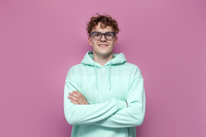 Jeune étudiant bouclé en hoodie à la menthe et lunettes se tient les bras croisés sur fond rose isolé images stock