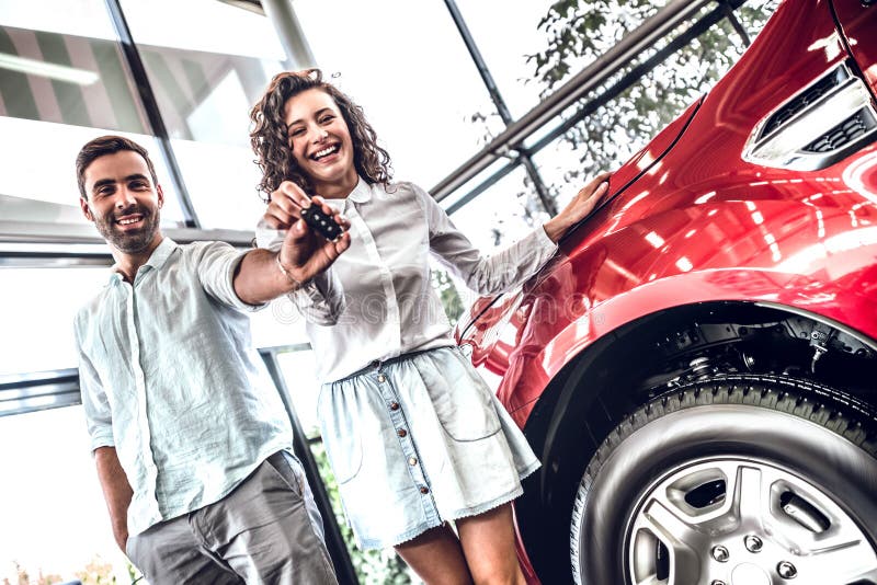 Jeunes beaux couples tenant la clé de voiture et regardant la caméra avec le sourire tout en se tenant à l'intérieur avec la voit images stock Jeunes beaux couples tenant la clé de voiture et regardant la caméra avec le sourire tout en se tenant à l'intérieur avec la voit images stock