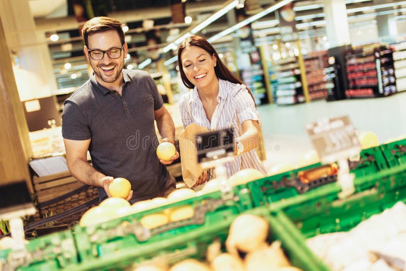 Jeunes couples affectueux gais dans le supermarché avec le chariot de achat choisissant des fruits photographie stock Jeunes couples affectueux gais dans le supermarché avec le chariot de achat choisissant des fruits photographie stock
