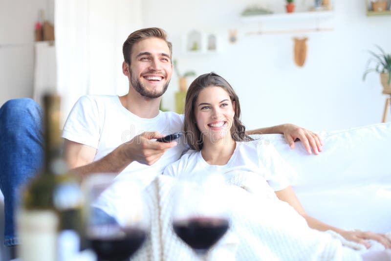 Jeunes couples affectueux sur le sofa à la maison regardant la TV et riant, buvant un verre de vin rouge images stock Jeunes couples affectueux sur le sofa à la maison regardant la TV et riant, buvant un verre de vin rouge images stock